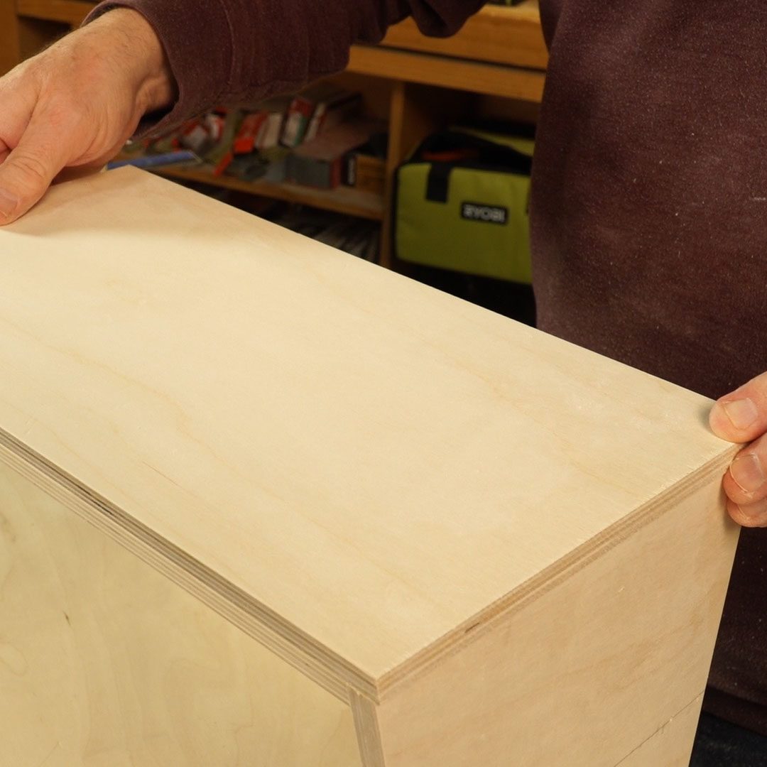 A person holds a light-colored plywood board, positioning it atop another similar piece in a workshop filled with tools and materials.