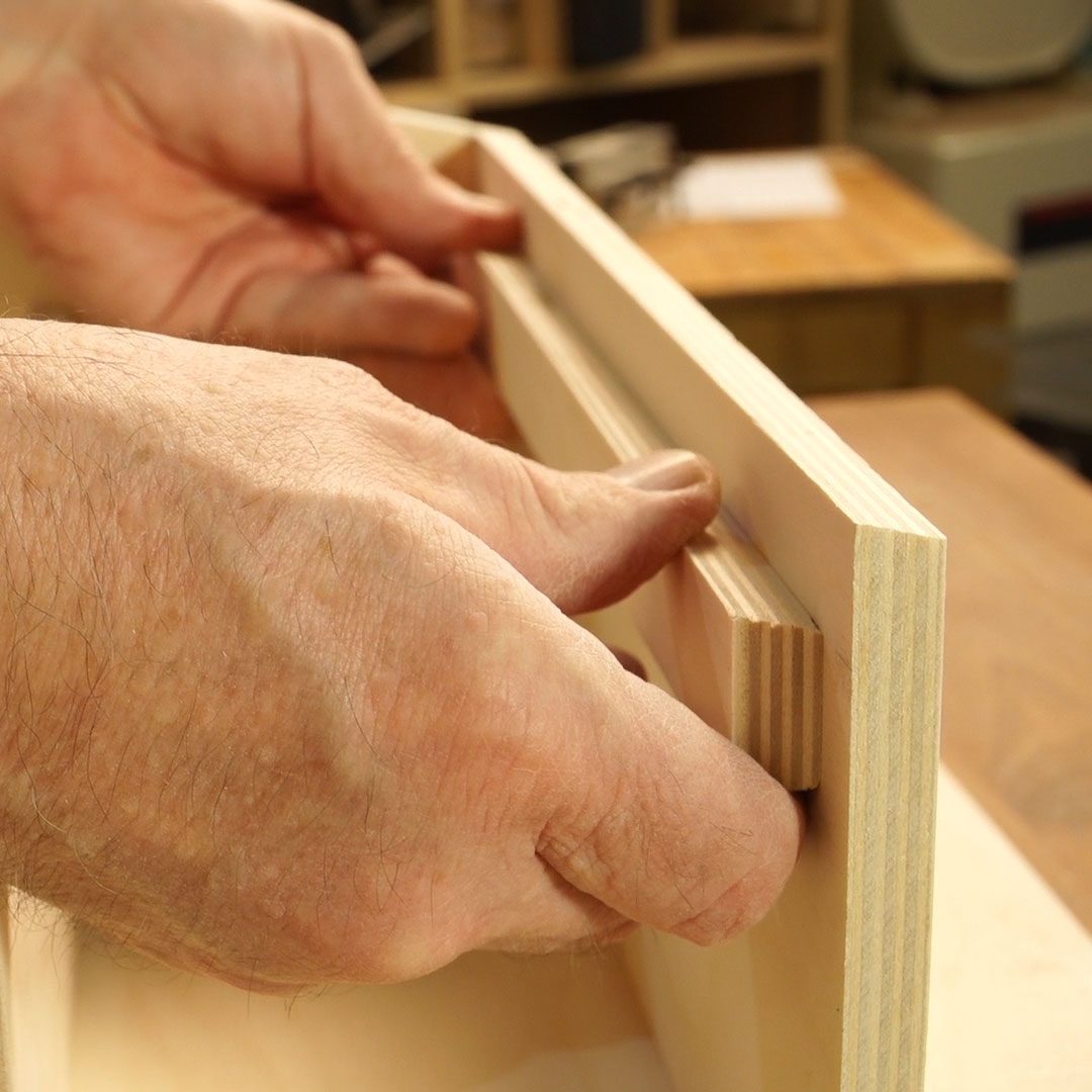 Hands adjust a wooden piece within a larger frame, focusing on precise alignment, in a woodworking shop setting with tools and materials in the background.