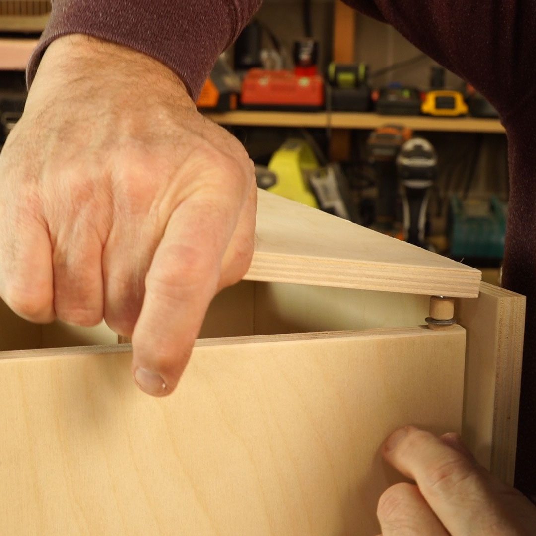 A hand is securing a wooden piece, aligning it with a connector, in a workshop filled with tools and equipment in the background.