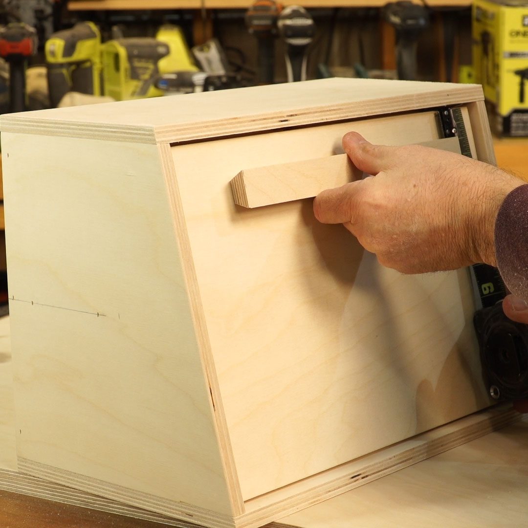 A hand pulls a wooden drawer handle from a light wooden box, situated in a workshop with tools visible in the background.
