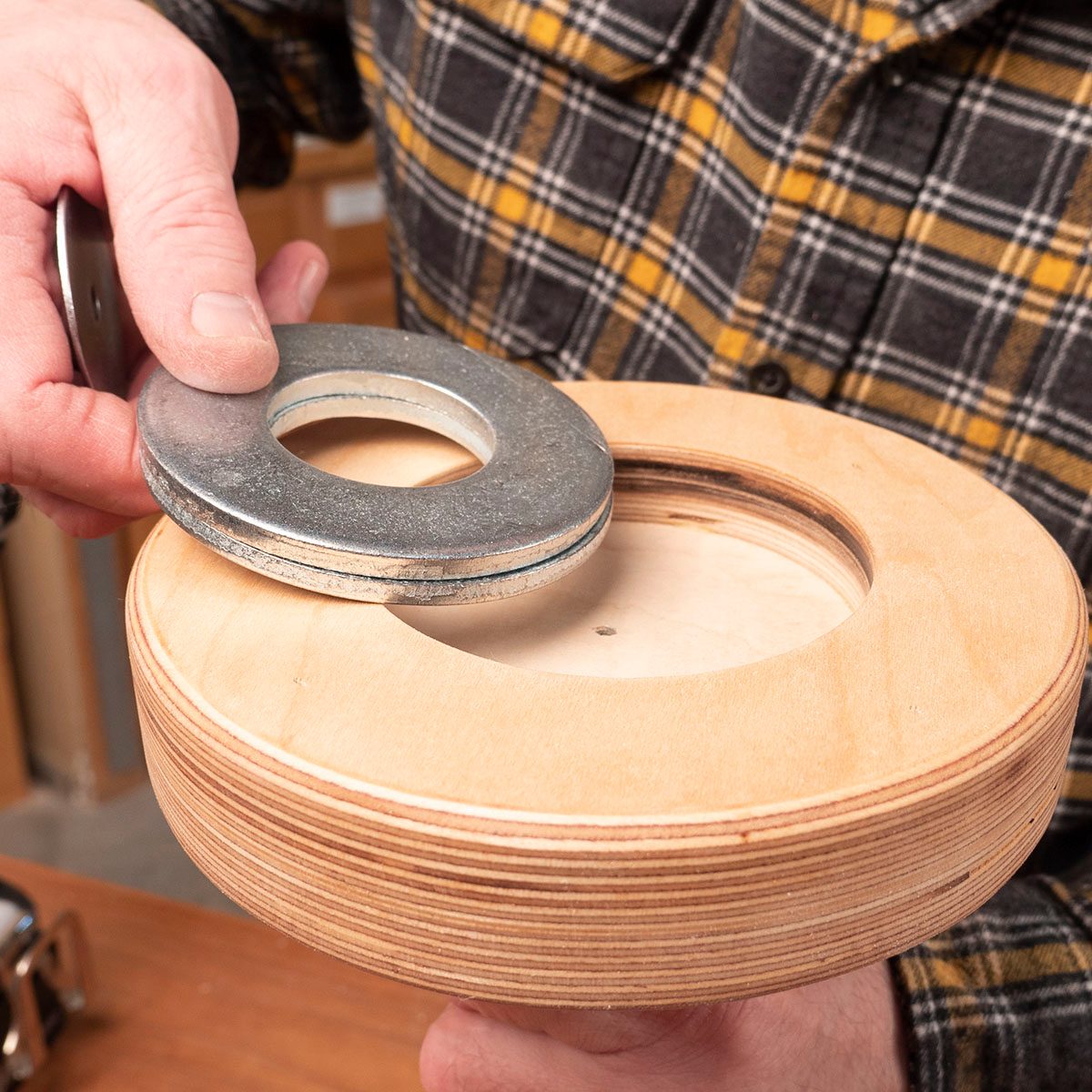 A hand holds a metallic washer above a wooden object with a circular cutout, positioned in a workshop setting with tools and materials nearby.