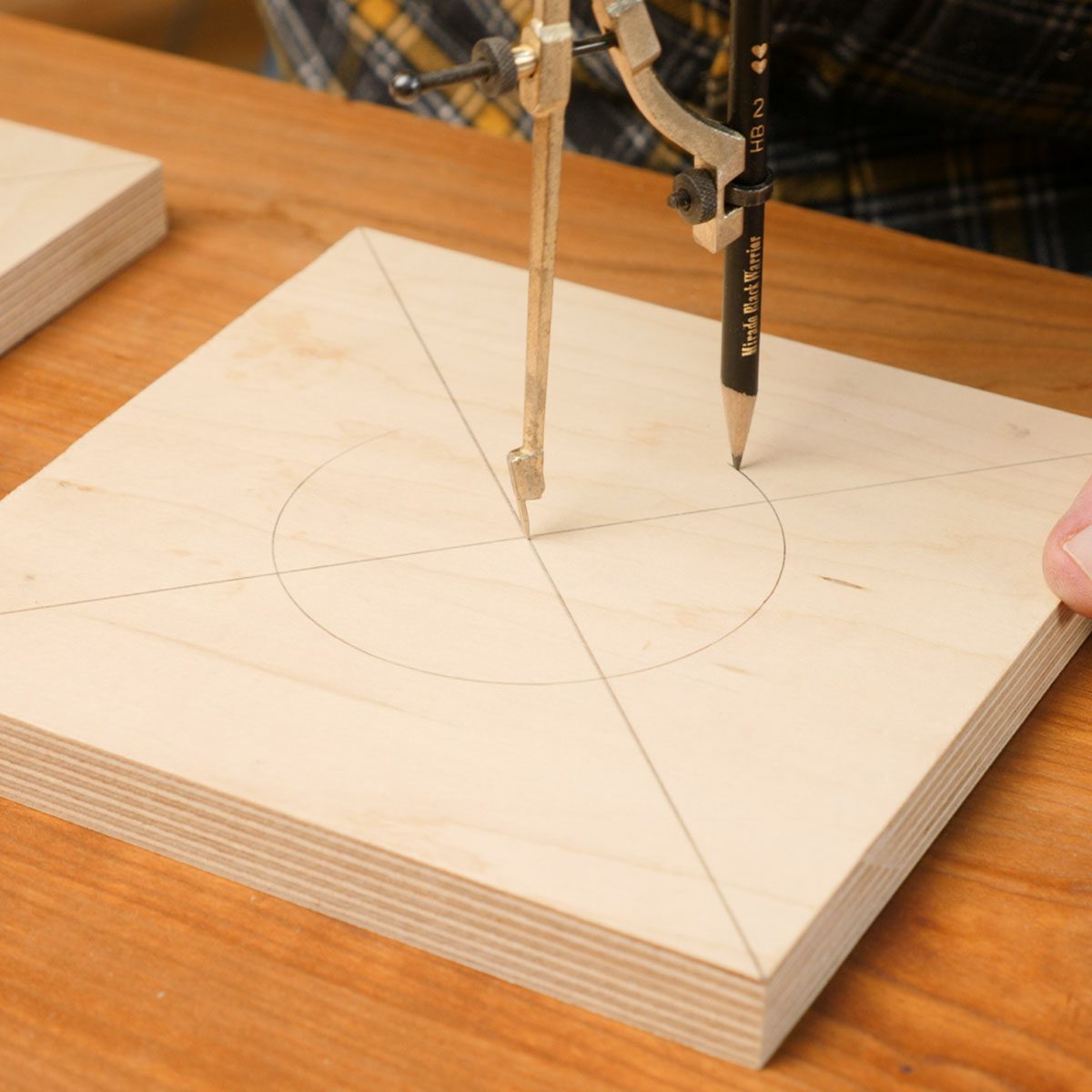 A compass draws a circle on a wooden square, which has diagonal lines marked, on a wooden table. Hands are visible guiding the compass.