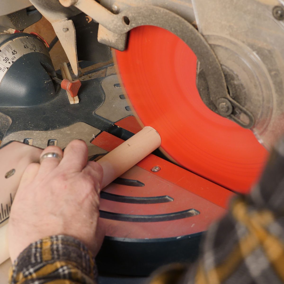 A hand holds a cylindrical object against a spinning red saw blade, preparing to cut it on a work surface filled with tools and equipment.