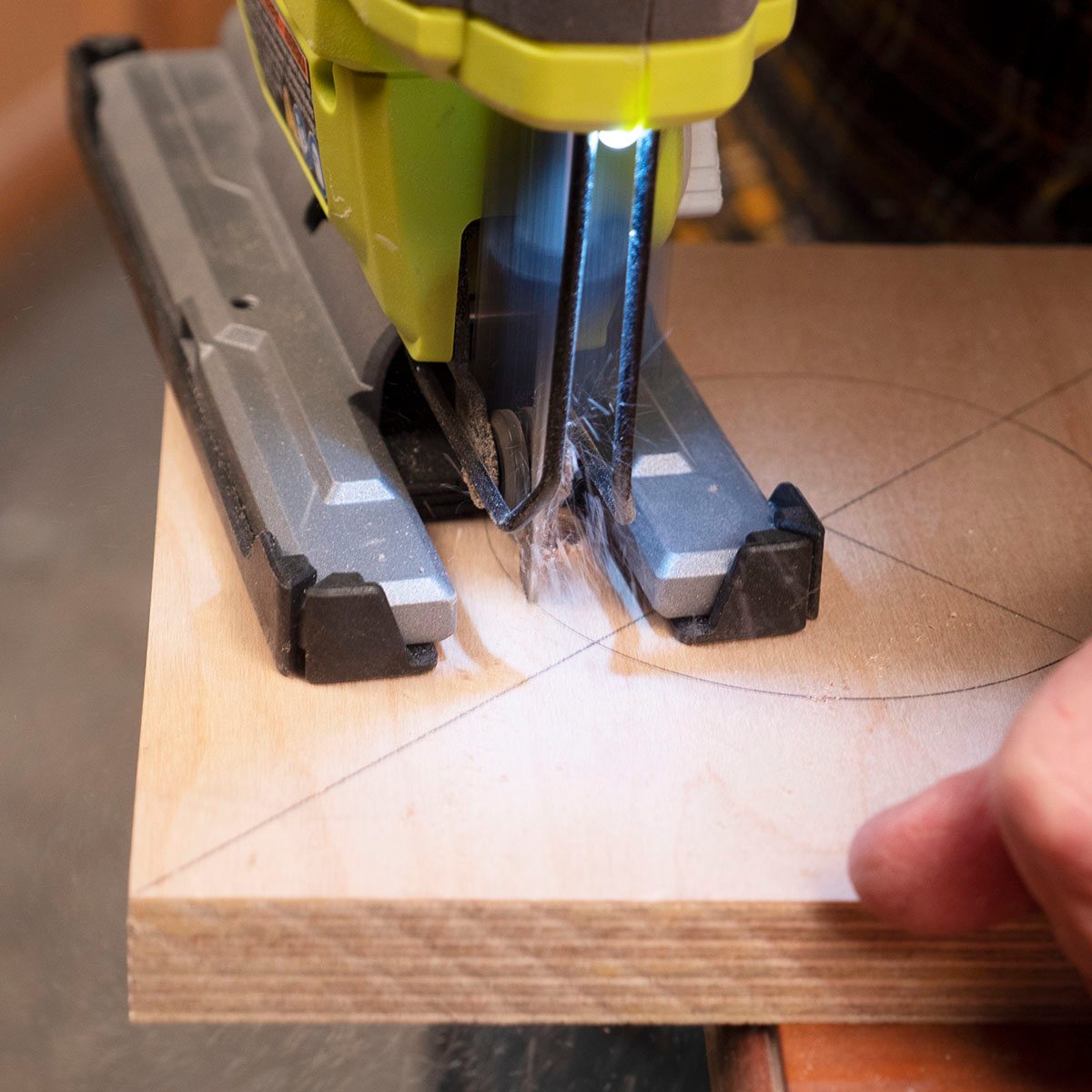A jigsaw cuts through a wooden board, with shavings flying, while a hand steadies the material nearby in a workshop setting.