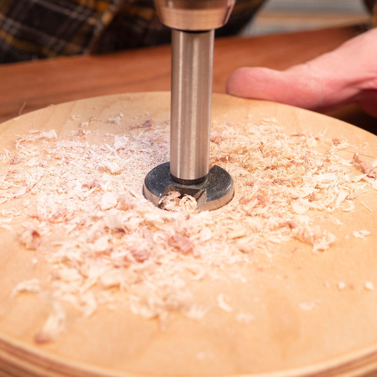 A drill bit is rotating above a wooden surface, creating shavings. The workspace appears to be a craft area with tools nearby.
