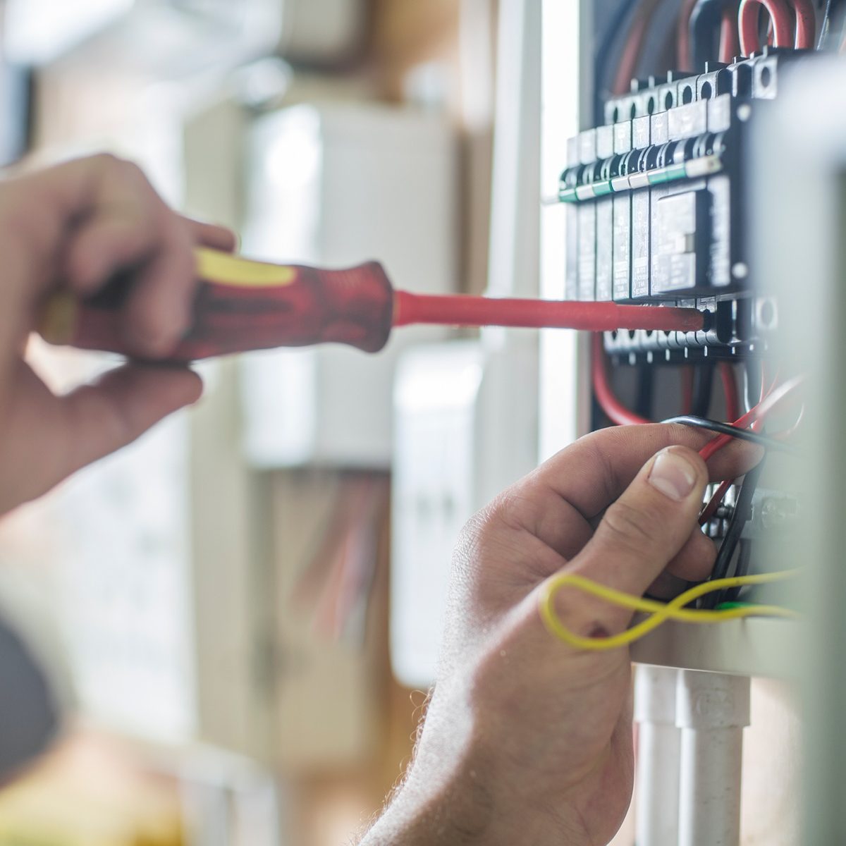 A hand uses a screwdriver to adjust wiring in an electrical panel, surrounded by various cables and devices in a workspace.
