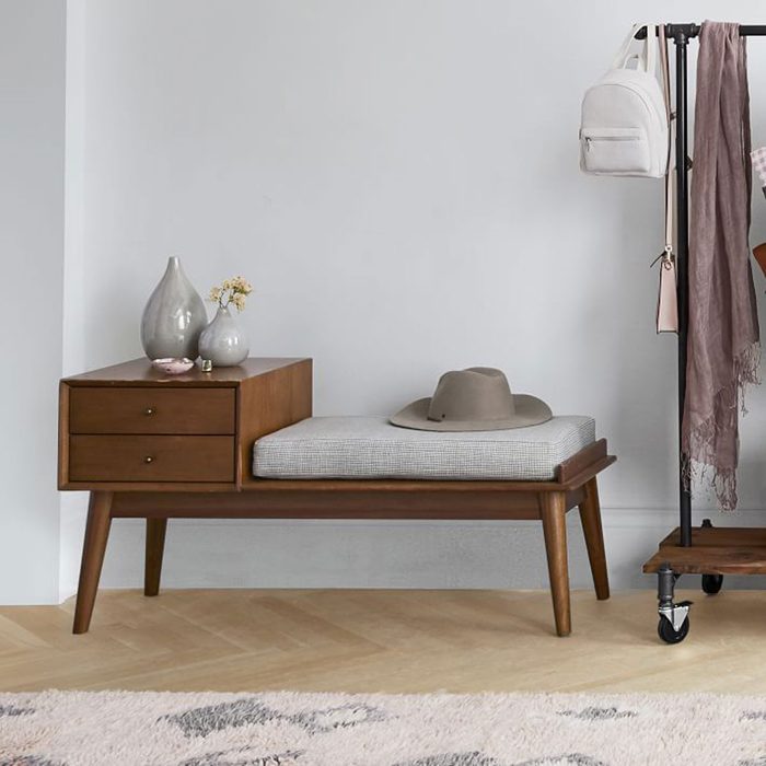 A wooden bench with a cushion displays a hat, accompanied by decorative vases and a rack holding bags and scarves in a minimalist room.