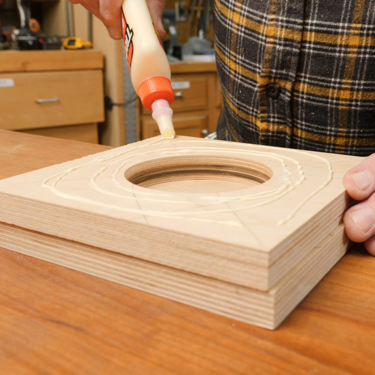 A person applies wood glue in a spiraling pattern on a wooden board with a circular cutout, positioned on a workbench in a workshop.