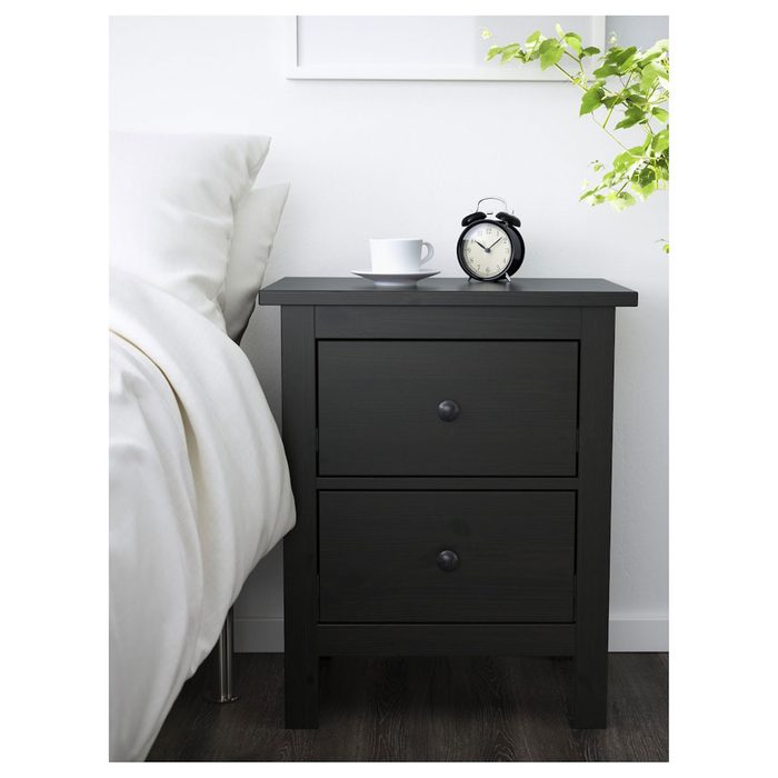 A black bedside table holds a cup and alarm clock, beside a white bed with fluffy pillows, in a light, minimal bedroom setting.