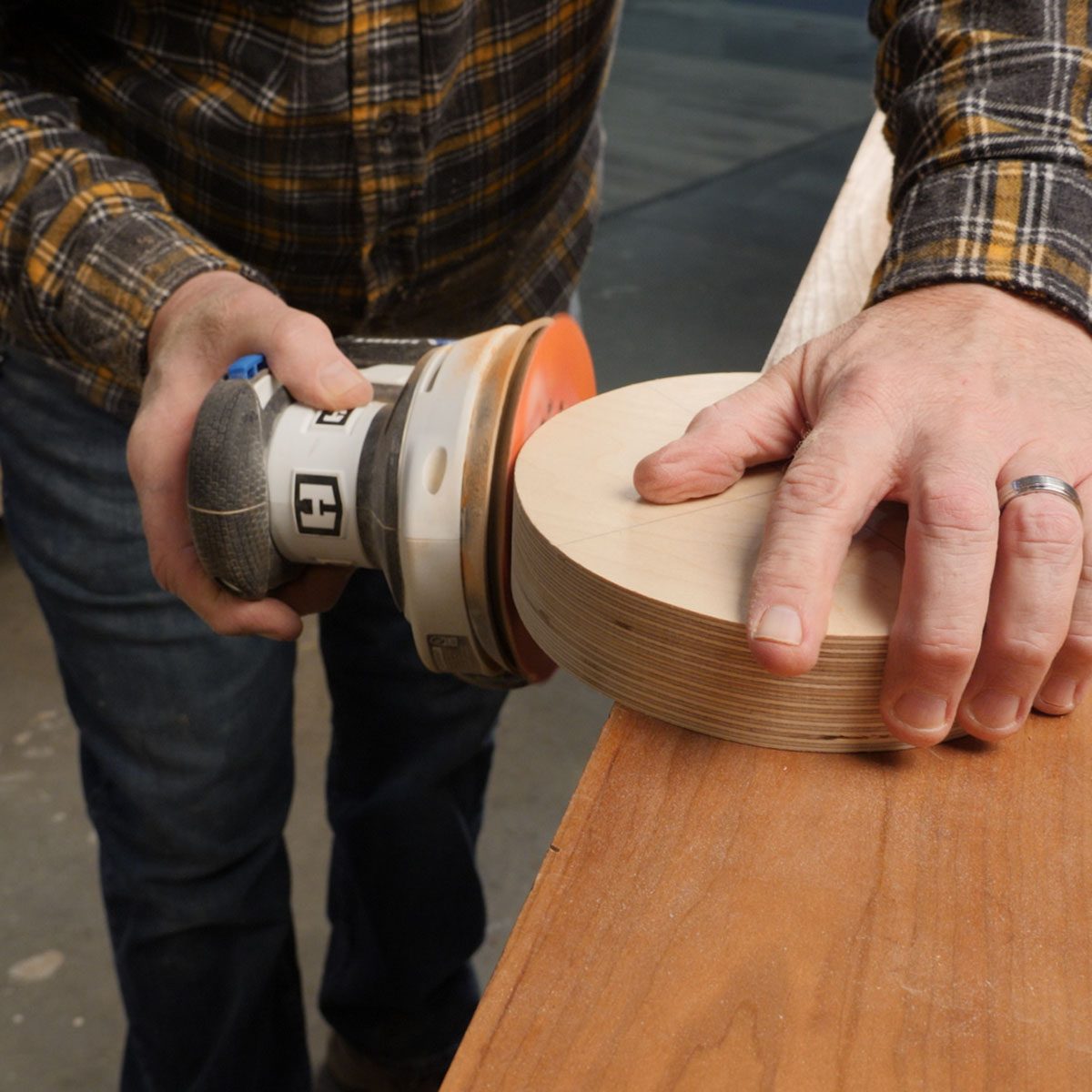 A person sands a circular wooden piece using an electric sander, with a workbench and a workshop backdrop indicating a crafting environment.