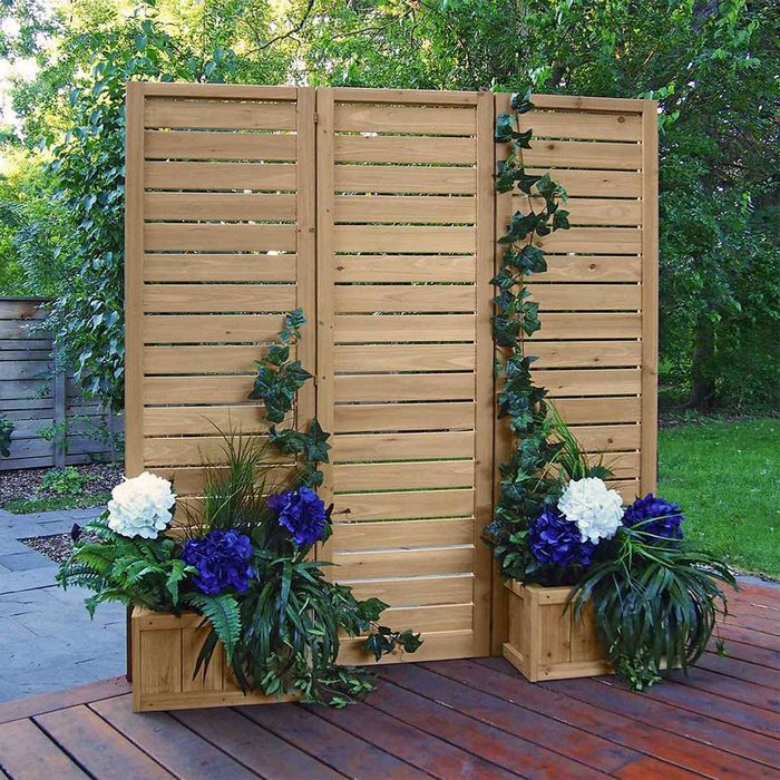A wooden privacy screen stands upright, adorned with greenery and flowers in planters, set against a serene garden backdrop with lush trees and greenery.