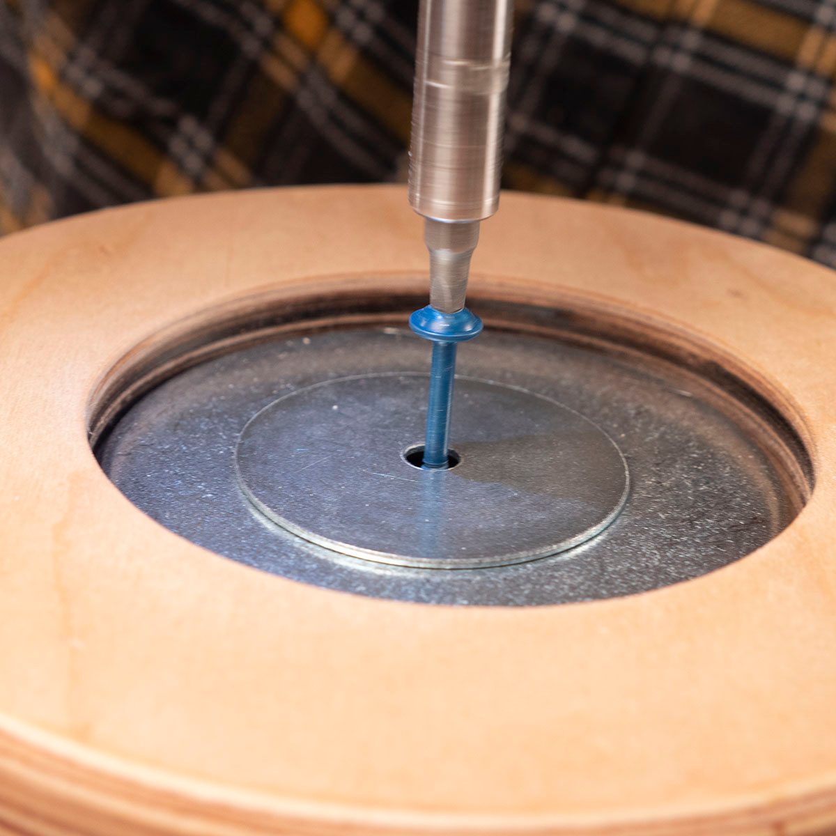 A drill bit is inserted into a metal disc, rotating above a wooden surface, as part of a crafting process in a workshop.
