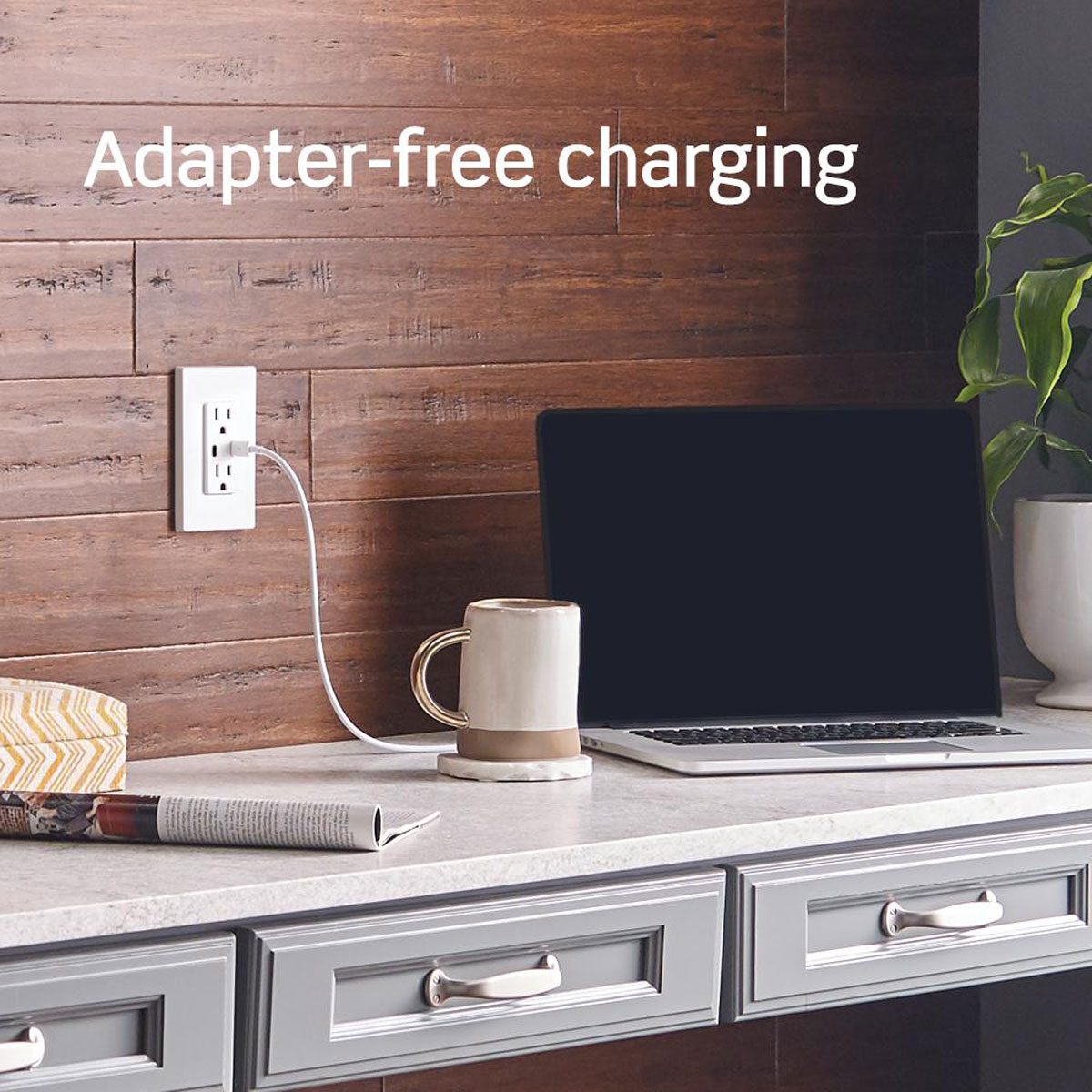 A laptop charges from a wall outlet, resting on a sleek countertop with a coffee mug, a decorative box, and a potted plant nearby.