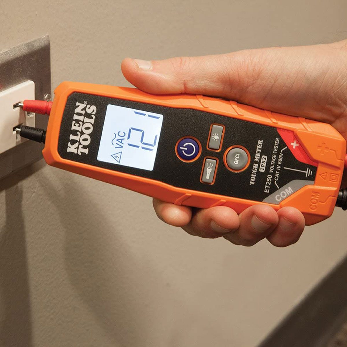 A hand holds a voltage tester, measuring voltage at a wall outlet, displaying "121" on its screen against a neutral-colored wall.