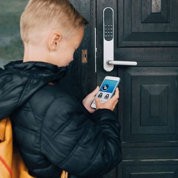 A child holds a smartphone to unlock a modern door lock, standing in front of a dark wooden door in an outdoor setting.