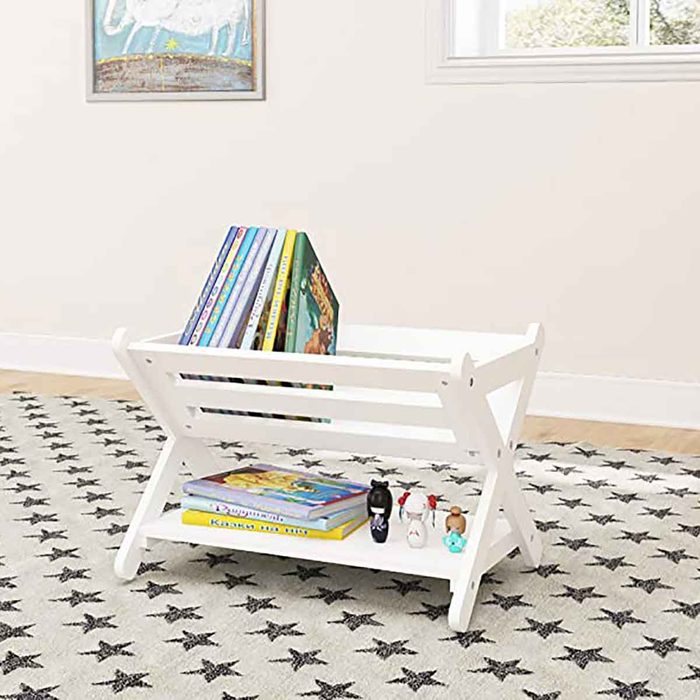 A white book rack holds colorful children's books and small toys, situated on a star-patterned rug in a bright, minimalistic room with a window.