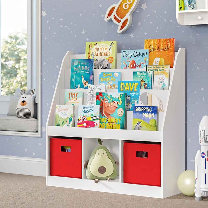 A white bookshelf holds a variety of colorful children's books, flanked by red storage bins. A plush avocado toy sits in one bin, with a cozy room backdrop.
