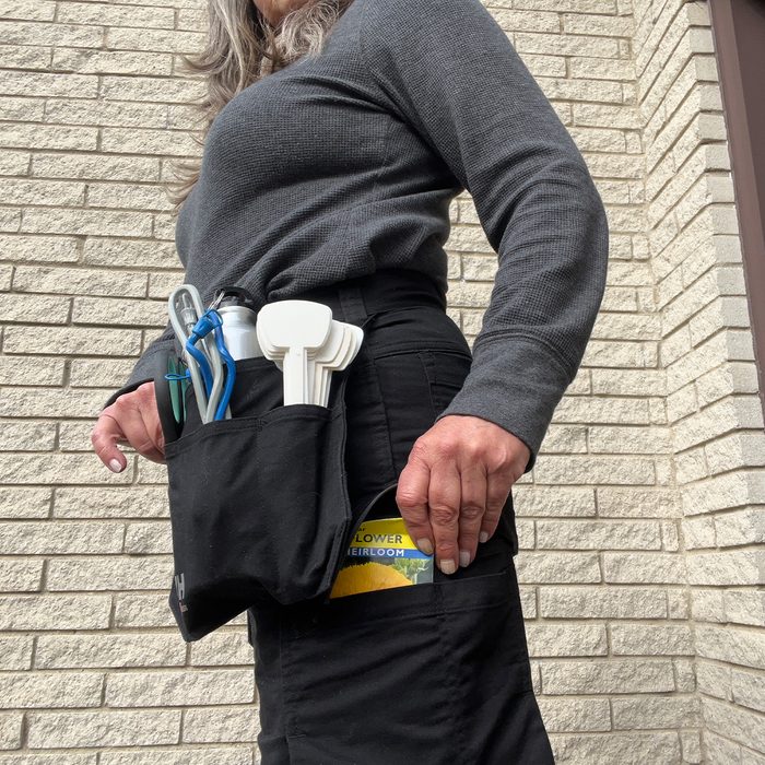 A person holds a gardening tool pouch with various tools and a seed packet, standing against a textured brick wall.