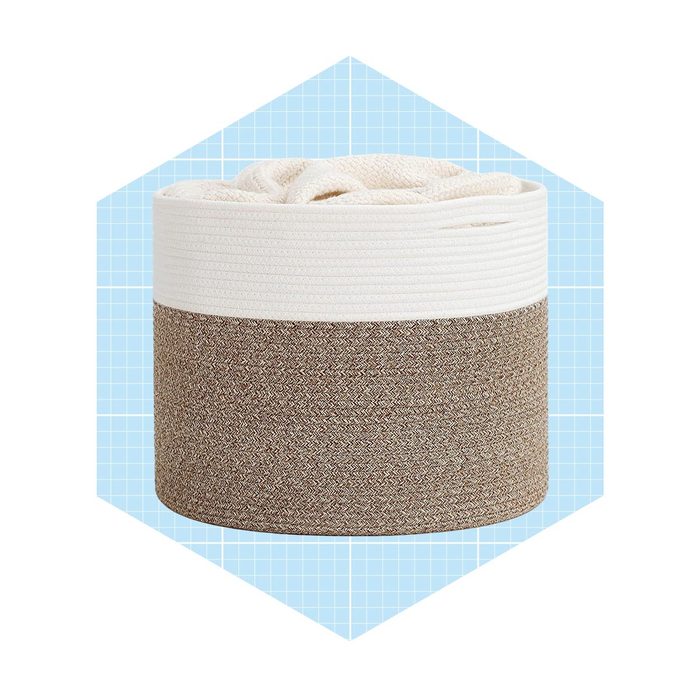 A cylindrical storage basket, filled with fabric, sits centered against a light blue grid background. The basket features a textured brown base and a white rim.