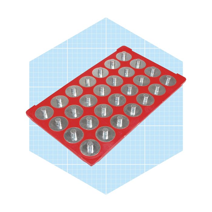A red silicone mold holds multiple circular depressions, each fitted with a small metal center. It's displayed on a light blue grid background.