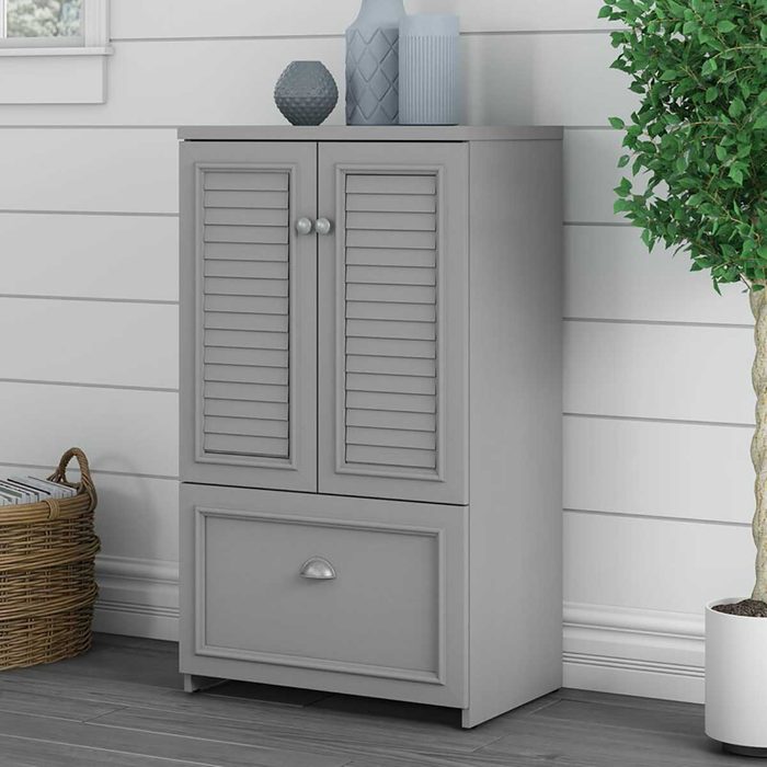 A gray cabinet with louvered doors and a drawer stands against a white shiplap wall, accompanied by decorative vases and a potted plant.