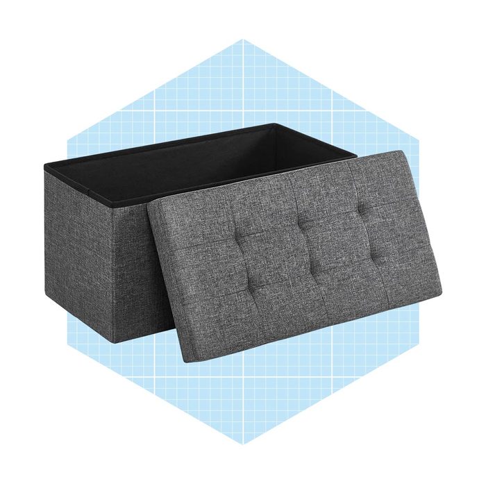A rectangular gray storage ottoman with a tufted lid is open, showcasing its interior. It's set against a light blue grid-patterned background.