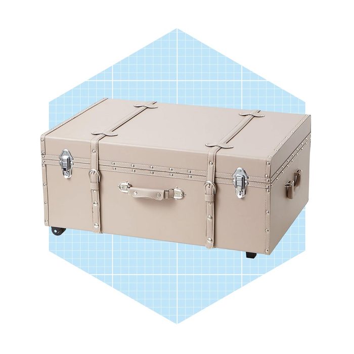 A beige trunk rests on wheels, featuring metal clasps and a handle, set against a light blue grid-patterned background.