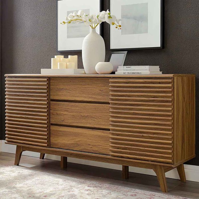 A wooden sideboard features slatted doors and three drawers, displaying decorative items, set against a textured wall with framed artwork.