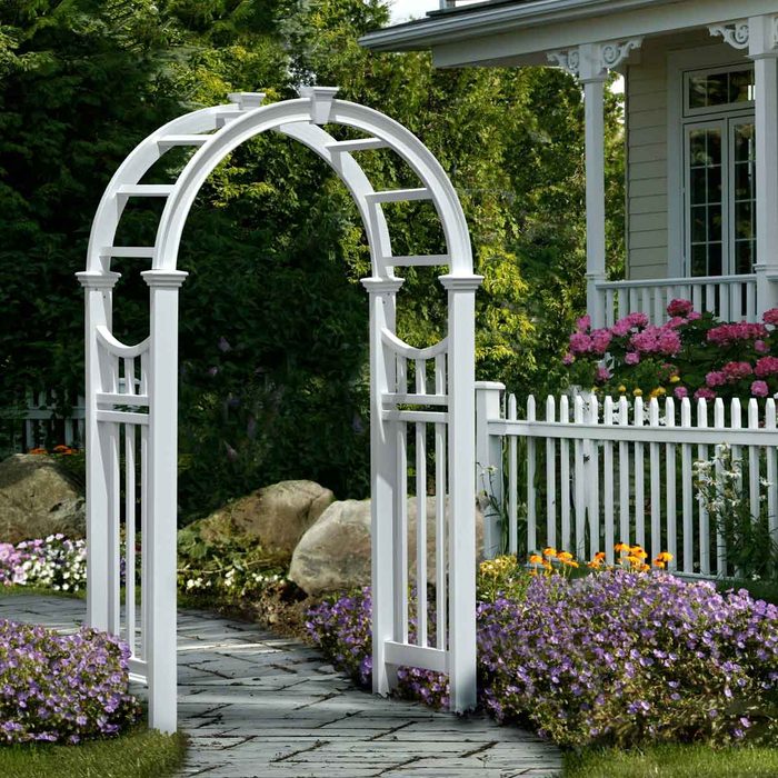 A white garden arch stands prominently along a stone path, surrounded by colorful flowers and lush greenery, leading to a house with a white fence.