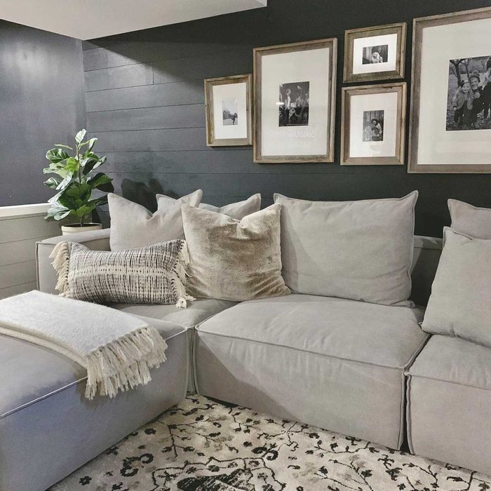 A sectional sofa with various cushions is positioned against a dark wall, surrounded by framed photos and a potted plant, on a decorative rug.
