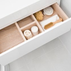A white drawer reveals neatly arranged bath accessories: sponges, a brush, a towel, and a container, set against a light tile floor.