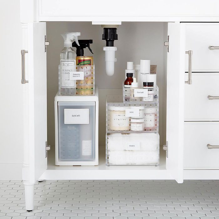 Bottles and containers are organized under a sink, holding cleaning supplies and skincare products, with white cabinetry and a hexagonal tiled floor surrounding them.
