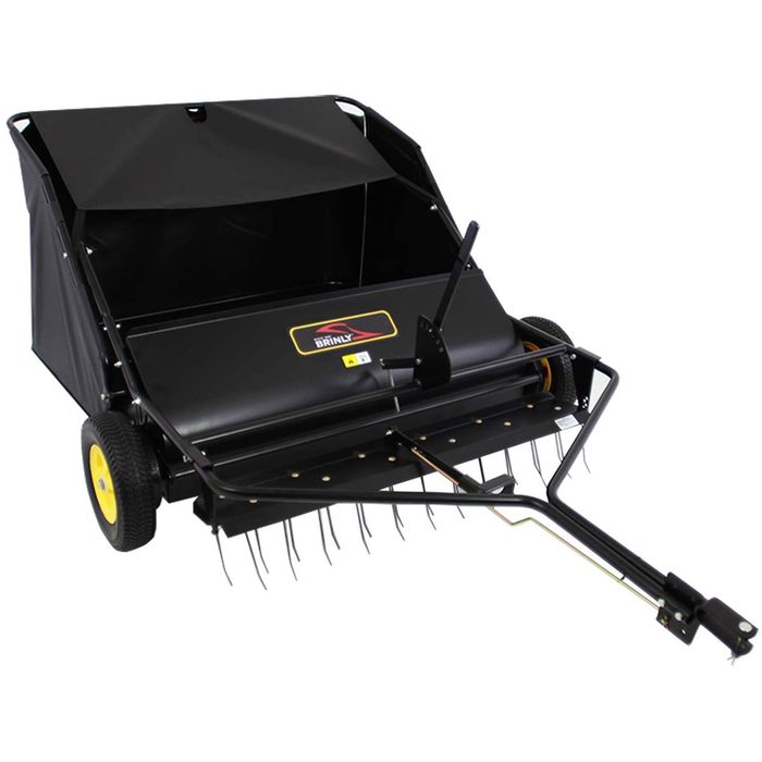 A tow-behind lawn sweeper collects debris using rotating brushes, with a black fabric collection bag, positioned on two yellow-rimmed wheels for easy maneuverability.