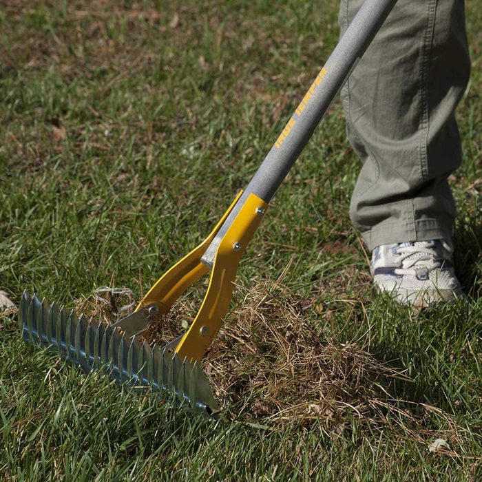 A gardening rake is removing debris from grass. A person stands nearby wearing light-colored pants and sneakers, focused on clearing the area.