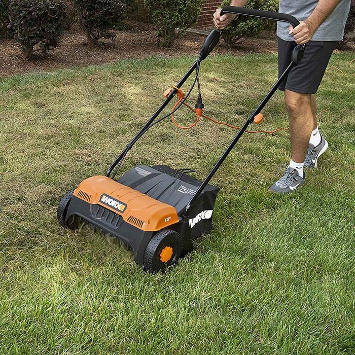 A powered lawn dethatcher moves over grass, removing thatch while a person holds the handle, demonstrating lawn care in a landscaped yard.