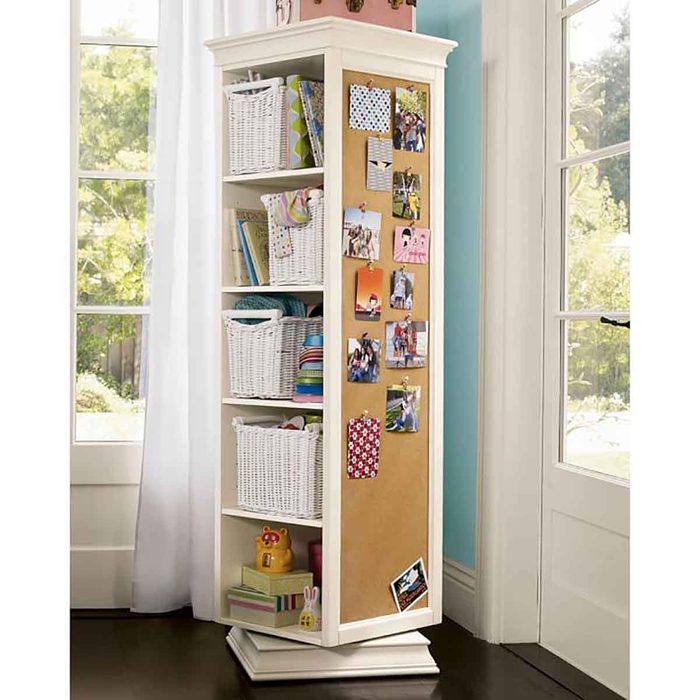 A multi-shelf storage unit displays wicker baskets and photo collages, standing in a well-lit room with large windows and light-colored walls.