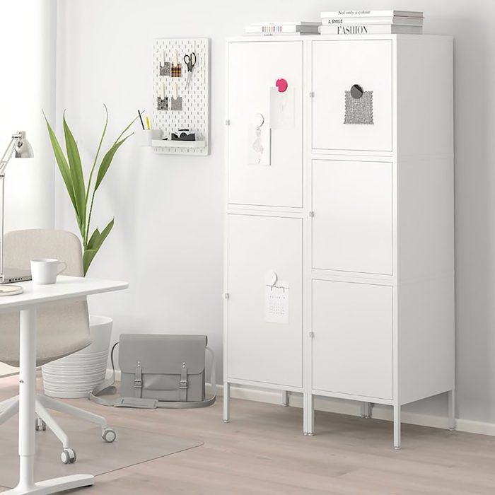 A white storage cabinet stands against a wall, with notes attached. A plant and a desk with a chair are nearby, creating a neat workspace.