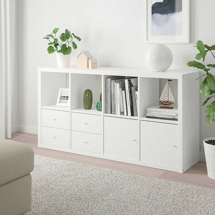 A white shelving unit holds books, decorative plants, and a model sailboat, positioned in a cozy room with light-colored furniture and soft carpeting.