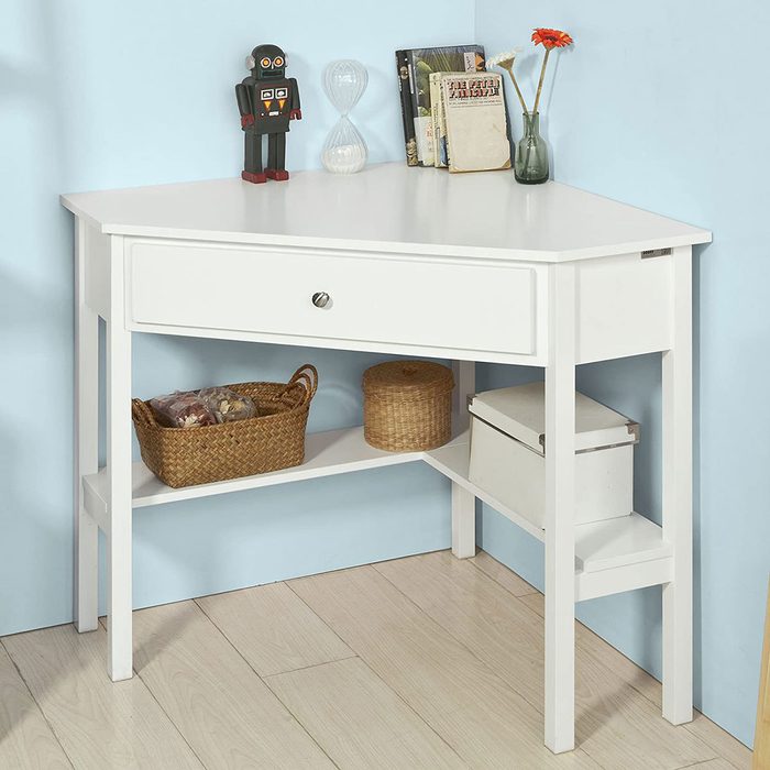 A white corner desk displays a toy robot and an hourglass. Below, woven baskets and a box are organized on shelves against a light blue wall.
