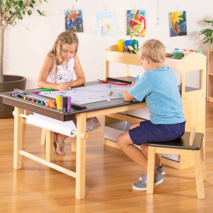 Two children draw on a table. One girl colors while a boy writes nearby. The room features artworks displayed and art supplies available.