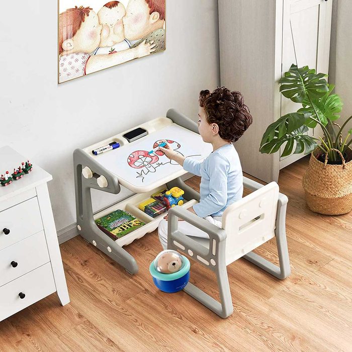 A child draws on a table while sitting in a chair, surrounded by toys and a plant, with a colorful painting hanging on the wall.