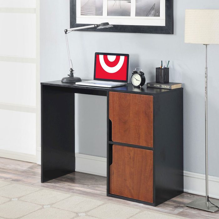 A desk with a laptop, lamp, clock, and stationery is positioned against a light-colored wall, accompanied by a standing lamp and a patterned rug.
