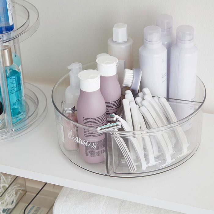 A clear round container holds various skincare and grooming products, including cleansers, razors, and brushes, organized on a light-colored shelf.