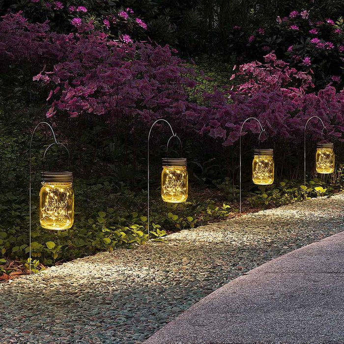 Mason jars filled with warm lights hang along a stone pathway, illuminating the surrounding purple flowers and greenery in a tranquil garden setting.