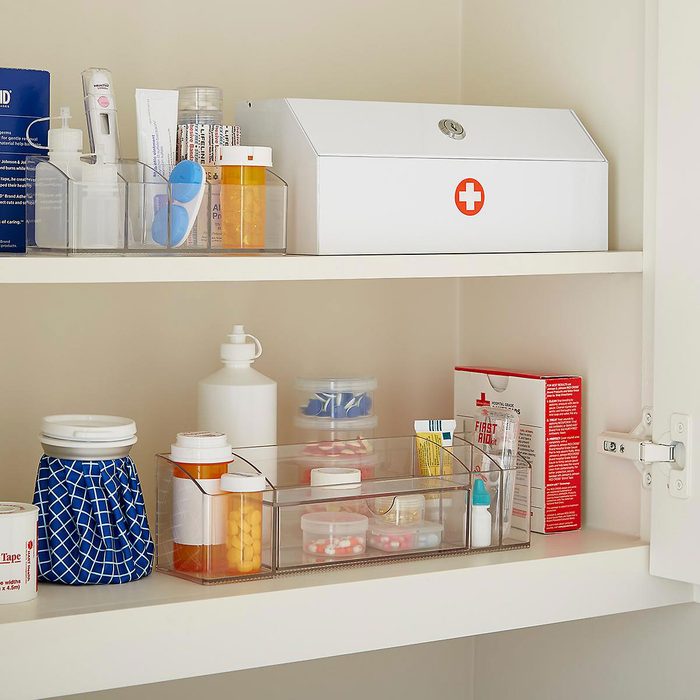 A medicine cabinet displays organized containers holding medications and medical supplies, with a first aid box and a thermometer, set in a neutral-colored room.