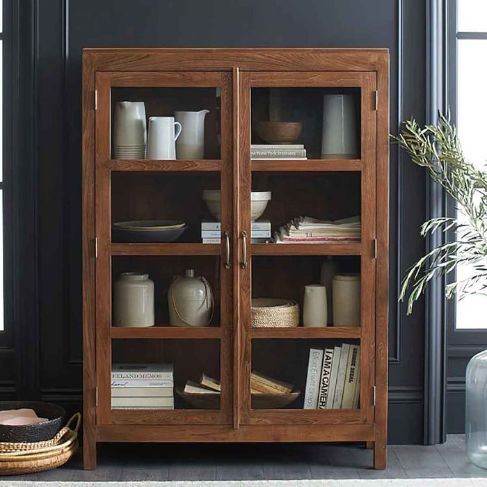 A wooden cabinet displays various dishes and books behind glass doors, situated against a dark wall with natural light filtering through nearby windows.