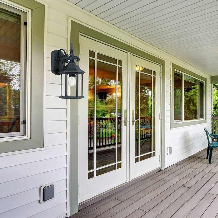 A black lantern is mounted beside glass double doors, illuminating a porch with wooden flooring, surrounded by greenery and glimpses of outdoor furniture.