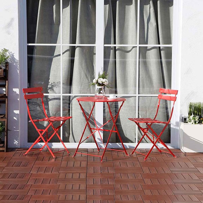 Red metal chairs and a round table sit on a wooden patio, adorned with a vase and coffee cup, in front of large windows with curtains.
