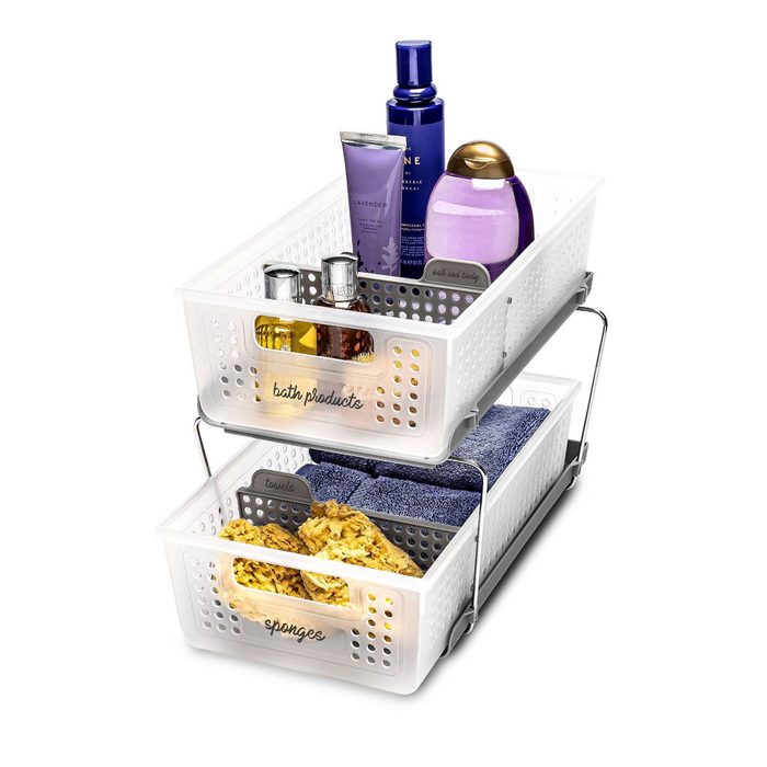 Two stacked storage baskets hold various bath products and towels. The top contains bottles, while the bottom has sponges and a folded towel.