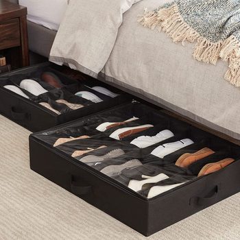 Two storage boxes beneath a bed hold various types of shoes, organized in compartments, surrounded by a light-colored rug and a textured bedding setup.